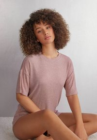 Ljusrosa ribbad t-shirt med korta ärmar, med en rund halsringning och avslappnad passform, bärs av en person som sitter på en ljus yta.