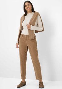 Femme portant un pull côtelé beige, un pantalon camel, des mocassins imprimé léopard et un pull camel drapé sur les épaules, se tenant contre un fond blanc.