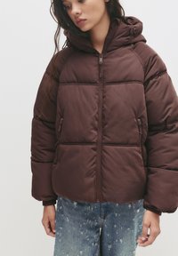 Braune Pufferjacke mit Kapuze, ausgestattet mit einem Reißverschluss und Seitentaschen. Weicher, gesteppten Textur mit einem cropped Design.