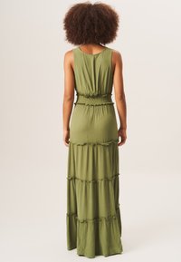 Olive green maxi dress with a tiered design, fitted waist, elastic smocking at the bodice, and a relaxed silhouette. Soft, textured fabric.