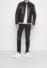 Zwarte leren jas met ritssluiting, wit T-shirt eronder, zwarte gescheurde jeans en witte sneakers met marineblauwe accenten.