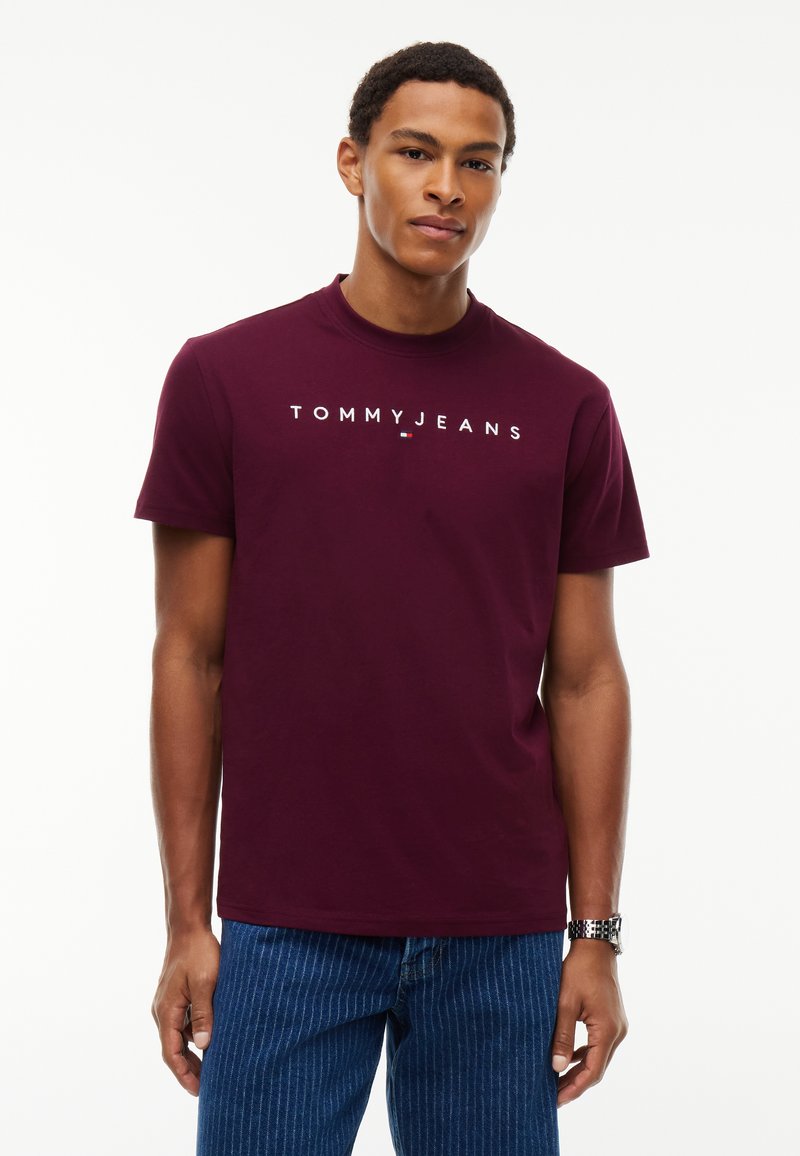 Νέος άνδρας που φοράει ένα μοβ Tommy Jeans t-shirt και μπλε τζιν με λεπτές ρίγες, στέκεται μπροστά σε ένα απλό λευκό φόντο.