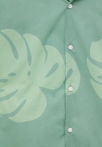 Camisa verde hecha de tela con un gran patrón de hojas en un verde más claro, con botones blancos a lo largo de la tapeta.