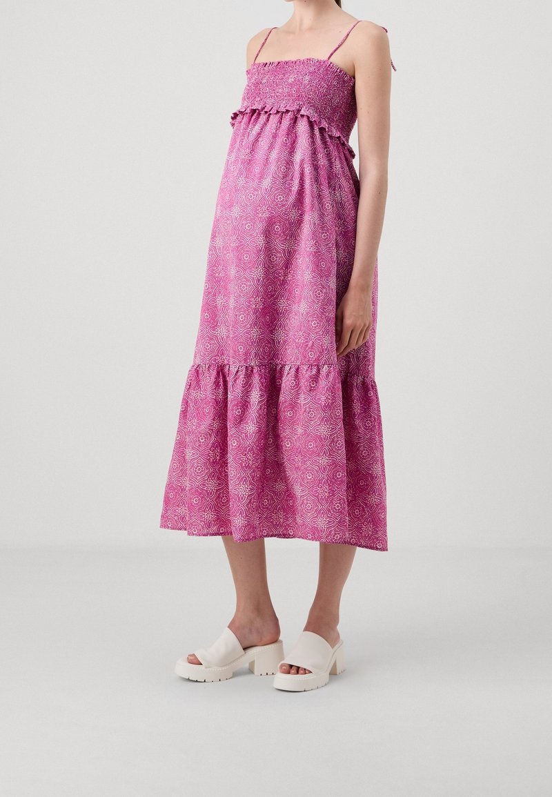 Femme portant une robe midi rose à motifs sans manches avec fines bretelles et des sandales blanches à talons blocs et orteils ouverts, debout sur un fond uni.