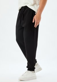 Schwarze Jogginghose aus weichem Stoff mit einem elastischen Bund, Seitentaschen und schmalen Bündchen. Getragen mit weißen Sneakern.