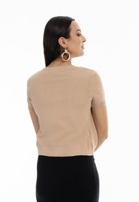 Beige svetr s krátkým rukávem a ribovanými manžetami a lemem, s hladkou texturou a přiléhavým střihem. Model nosí velké kruhové náušnice.