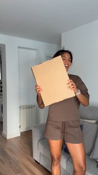 Cutie de carton maro ținută vertical, având o suprafață plană și netedă cu colțuri decupate, plasată pe un fundal neutru cu pardoseală din lemn.