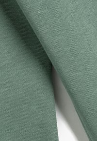 Tejido de punto en color verde apagado, con textura suave y finas rayas. Dos capas superpuestas, mostrando un material suave y transpirable.
