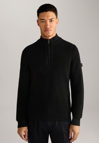 Schwarzer gerippter Strickpullover mit hohem Kragen und Viertelreißverschluss, ausgestattet mit langen Ärmeln und einem seitlichen Etikett. Glatte Textur über die gesamte Strecke.