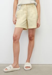 Beige bomullsshorts med avslappnad passform, utrustade med sidosfickor och en mjuk textur. Matchas med vita fluffiga slip-insandaler.
