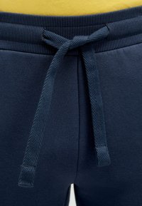 Nahaufnahme von marineblauen Jogginghosen mit einem gebundenen Kordelzugbund und einem Hauch eines gelben Shirts darüber.