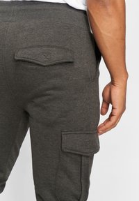 Szare jogger pants wykonane z miękkiego materiału z prążkowanym paskiem w talii, z dwoma kieszeniami zapinanymi na klapę z tyłu oraz jedną kieszenią cargo z boku.