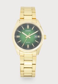 Montre Casio ton or avec un cadran dégradé vert, des index argentés et un affichage de date. Elle possède un bracelet en métal doré à maillons et une lunette texturée.