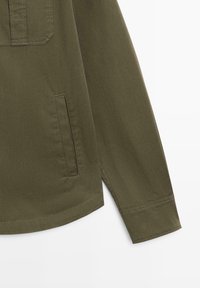 Dettaglio di una camicia verde oliva a maniche lunghe con una tasca laterale, cuciture visibili all'orlo e polsino risvoltato su uno sfondo uniforme.