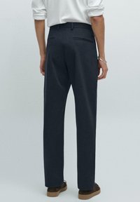 Tmavě šedé kalhoty s rovnými nohavicemi, dvěma zadními kapsami a hladkým povrchem, v kombinaci s hnědými botami, pohled zezadu.