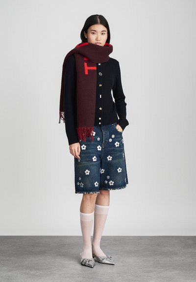 Cardigan noir tricoté avec des boutons dorés, jupe en denim brodée de fleurs, écharpe bordeaux avec des accents rouges, chaussures argentées et chaussettes blanches transparentes.