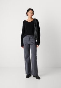 Egy nő fekete kötött pulóvert, szürke széles szárú nadrágot és fekete cipőt visel, miközben egy fekete pliszírozott táskát tart a vállán.