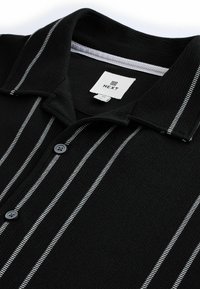 Camisa negra de cuello con rayas verticales blancas, tejido texturizado y una botonera. Presenta una etiqueta marcada "NEXT" en el interior del cuello.