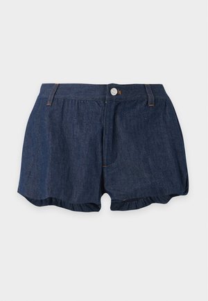 Femme debout portant un polo bleu clair à manches courtes, un short en jean foncé et des sandales à talons noirs sur fond blanc.