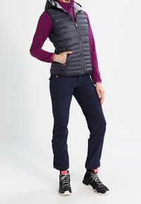 Gepolsterte, ärmellose Weste in Dunkelgrau mit Reißverschluss vorne und Kapuze, kombiniert mit blauen Hosen. Schwarze Wanderschuhe und pinke Knöchelsocken.