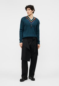 Homme aux cheveux bouclés portant un pull en maille torsadée bleu sarcelle avec des rayures orange et bleu marine, un pantalon large noir avec un détail noué, et des chaussures noires.