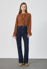 Brun bluse med volanger og knyting ved halsen, sammen med blå slengbukser. Modellen har på seg brune hæler. Bakgrunnen er nøytral.