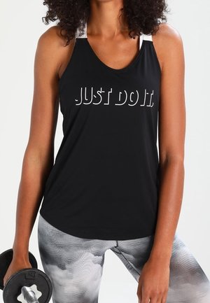 Femme portant un débardeur noir avec le texte "Just Do It", tenant un haltère dans une main, vêtue de leggings gris à motifs.