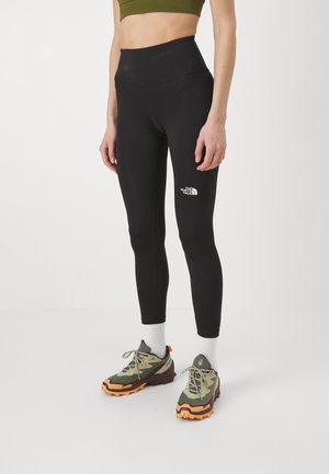 Leggings noirs en tissu extensible, design taille haute, avec un logo blanc sur la cuisse. Portés avec des chaussures vert olive et orange.