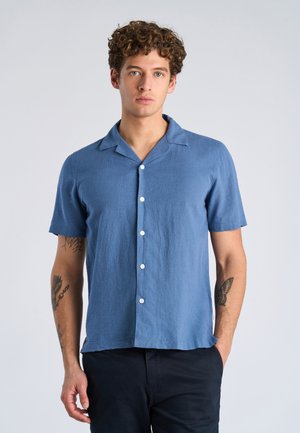 Jeune homme aux cheveux bouclés portant une chemise bleue à manches courtes boutonnée et un pantalon noir, debout devant un fond clair uni.