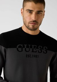 Schwarz-grauer Pullover mit einem auffälligen "GUESS"-Logo und dem Schriftzug "EST. 1981". Gestrickte Textur mit rundem Ausschnitt und langen Ärmeln.
