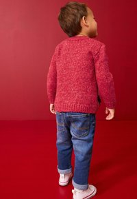 Pull en tricot rouge avec un motif texturé, associé à un jean bleu et des chaussures blanches. L'enfant est de dos, montrant la vue arrière.