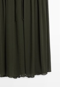 Robe en tissu léger vert foncé avec de fines plis verticaux, nouée à la taille avec des cordons assortis, ourlet légèrement courbé.