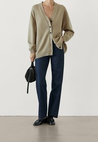 Beige knit cardigan with a V-neck, button front, and ribbed cuffs, paired with dark denim trousers and black shoes. Black bag in hand.