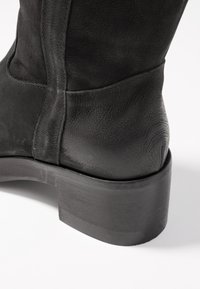 Czarny skórzany botek sięgający kostki z teksturowanym wykończeniem, okrągłym noskiem i masywnym obcasem. Posiada subtelne przeszycia wzdłuż szwów oraz gładką, niskoprofilową podeszwę.