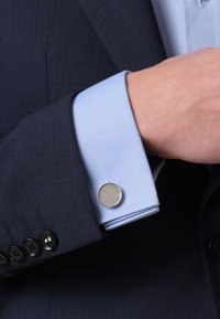 Marineblauer Sakko mit schwarzen Knöpfen, hellblauem Shirtärmeln, die mit einer runden, strukturierten silbernen Manschettenknopf verziert sind.