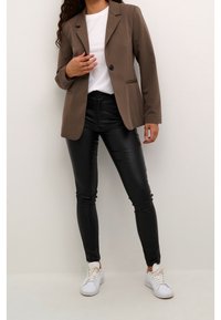 Blazer marron avec un bouton unique et des détails de poche, porté sur un t-shirt blanc. Associé à un pantalon noir en similicuir et des baskets blanches.