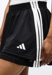 Κοντινό πλάνο προσώπου που φοράει μαύρο αθλητικό σορτς με τρεις λευκές ρίγες και λευκό λογότυπο κοντά στο τελείωμα, κρατώντας την άκρη του σορτς.