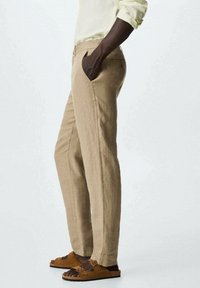 Pantalon en lin beige à coupe droite, poches latérales et texture subtile, associé à des sandales marron. Le tissu tombe élégamment.