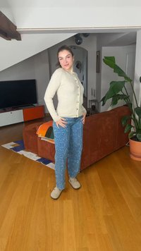 Woman standing in a living room wearing a beige buttoned cardigan, blue patterned jeans, and beige shoes, with a brown couch and plant nearby.