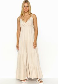 Jasno beżowa sukienka maxi z body w stylu kopertowym, warstwową spódnicą z falbanami i cienkimi ramiączkami. Nosi ją w połączeniu z białymi sandałami na cienkich paskach.