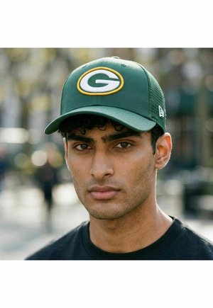 Mann mit lockigem schwarzem Haar, der eine grüne Kappe mit einem gelben und weißen "G"-Logo trägt, steht im Freien vor einem unscharfen städtischen Hintergrund.