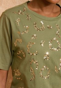 Olivgrünes T-Shirt aus weichem Stoff, das mit verstreuten goldenen Pailletten in verschiedenen Formen verziert ist, die ein Muster auf der Vorderseite bilden.