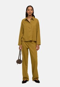 Koszula w paski w musztardowym kolorze z kołnierzykiem i kieszonką z przodu, zestawiona z dopasowanymi spodniami. Dodatki obejmują małą okrągłą brązową torbę i czarne buty.
