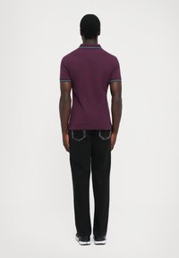 Άνδρας που στέκεται γυρισμένος προς τα πίσω, φορώντας μοβ polo μπλούζα με ριγέ περιθώριο, μαύρα τζιν και μαυρόασπρα αθλητικά παπούτσια, σε λευκό φόντο.