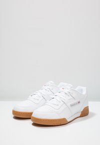 Sneaker sportive bianche con tomaia in pelle, punta perforata e suola in gomma naturale a contrasto. Presenta logo e design tradizionale con lacci.