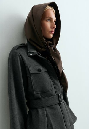 Woman wearing a dark grey belted coat and a brown hooded scarf, looking to the side against a light background.