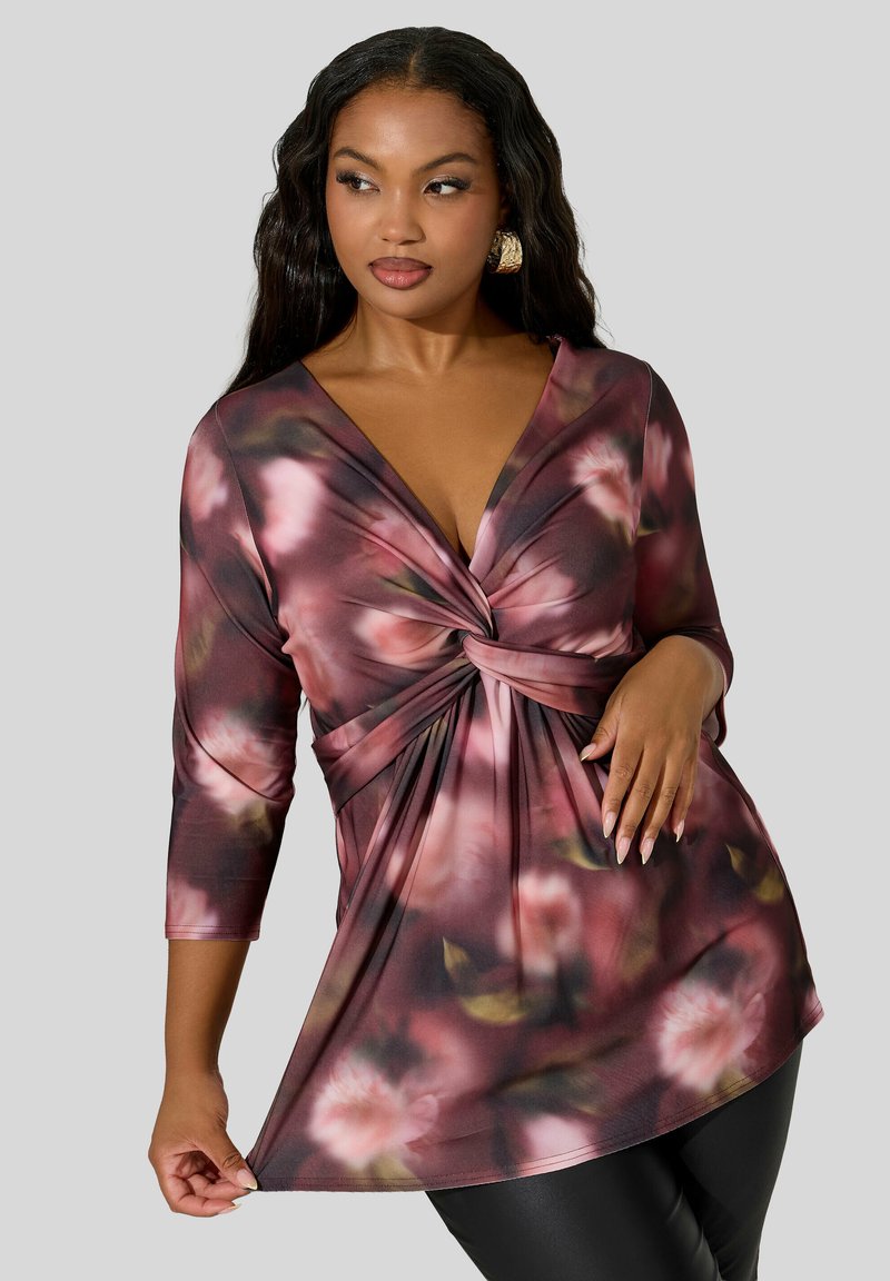 Vrouw in een bloemenpatroon, met een gedraaide voorkant, lange mouwen en een V-hals, gecombineerd met zwarte broek tegen een grijze achtergrond.