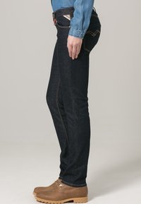 Jeans de mezclilla azul oscuro con un corte slim, con costuras doradas y una cinturilla de altura media. Complementados con una camisa de botones azul claro.