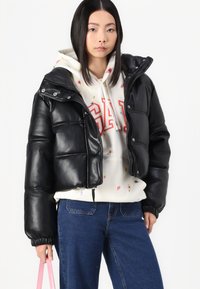 Zwarte cropped pufferjack gemaakt van leerachtig materiaal met een hoge kraag, ritssluiting en elastische manchetten. Gelaagd over een witte hoodie.