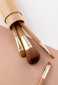 Bambus-Make-up-Pinsel mit goldfarbenen Ferrulen, ausgestattet mit weichen Borsten. Ein zylindrischer Bambushalter präsentiert die Pinsel auf einer strukturierten, hellbraunen Oberfläche.
